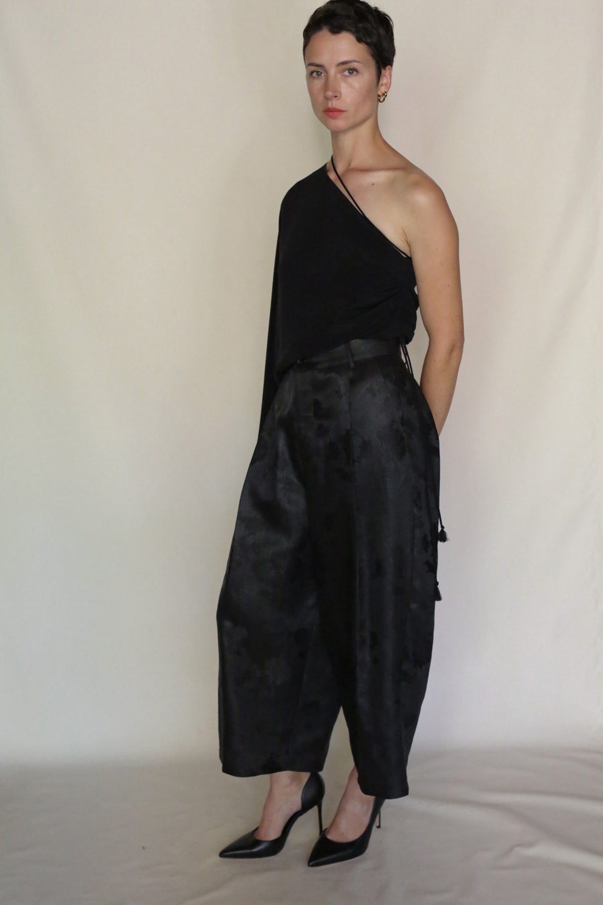 A person with short dark hair wears a sculptural cropped one-shoulder black top, Samurai Pants, and black high heels while standing against a plain light-colored background.