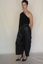 Load image into Gallery viewer, A person with short dark hair stands against a plain background, wearing a black one-shoulder top, black pointed high heels, and the Samurai Pants | Xiang Yun Silk Lingering Fragrance in high-waisted loose silk jacquard. Their hands are behind their back.