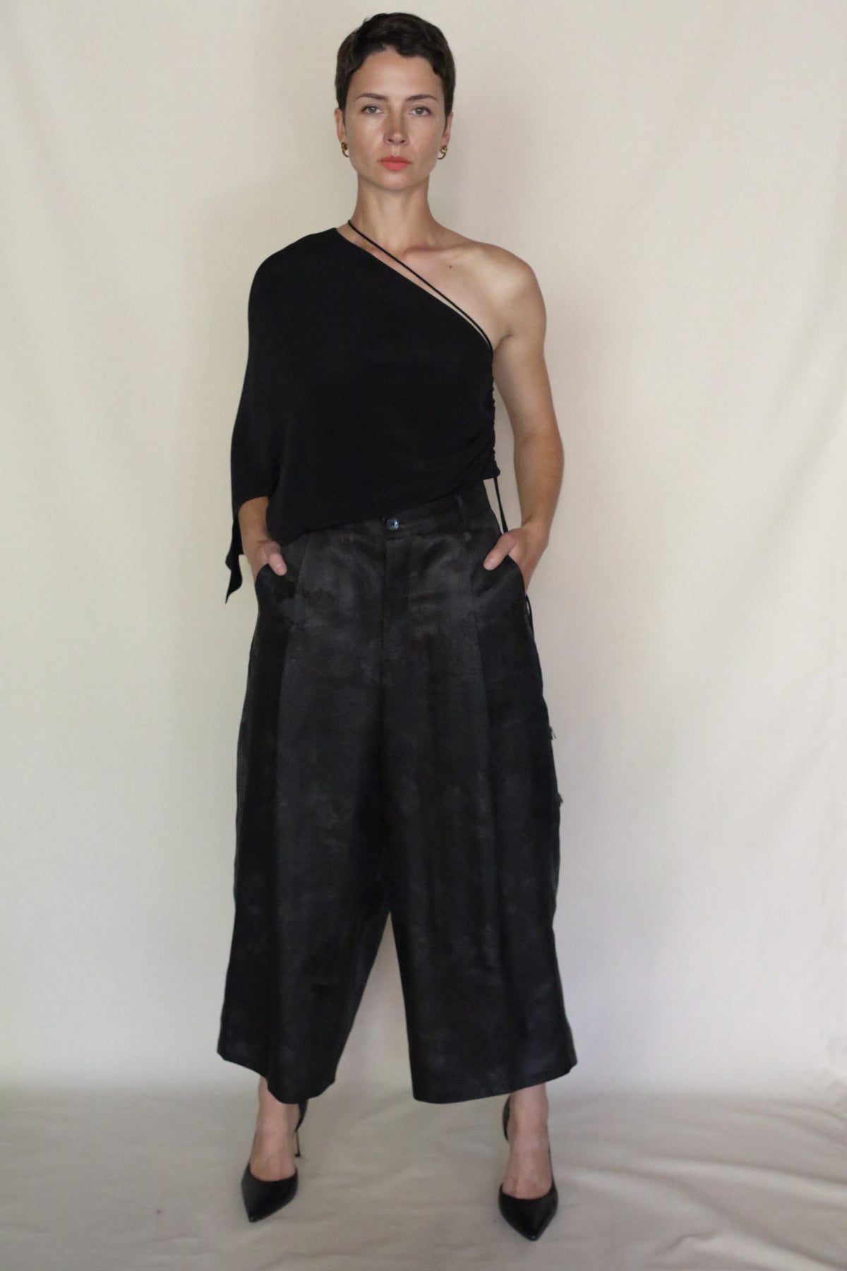 A person with short dark hair stands against a plain background, wearing a black one-shoulder top, Samurai Pants, and black heels, hands in pockets and a neutral expression.