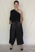 Load image into Gallery viewer, A person stands against a plain background wearing a black, one-shoulder top, Samurai Pants | Xiang Yun Silk Lingering Fragrance in silk jacquard, and black heels, hands in pockets with a neutral expression.