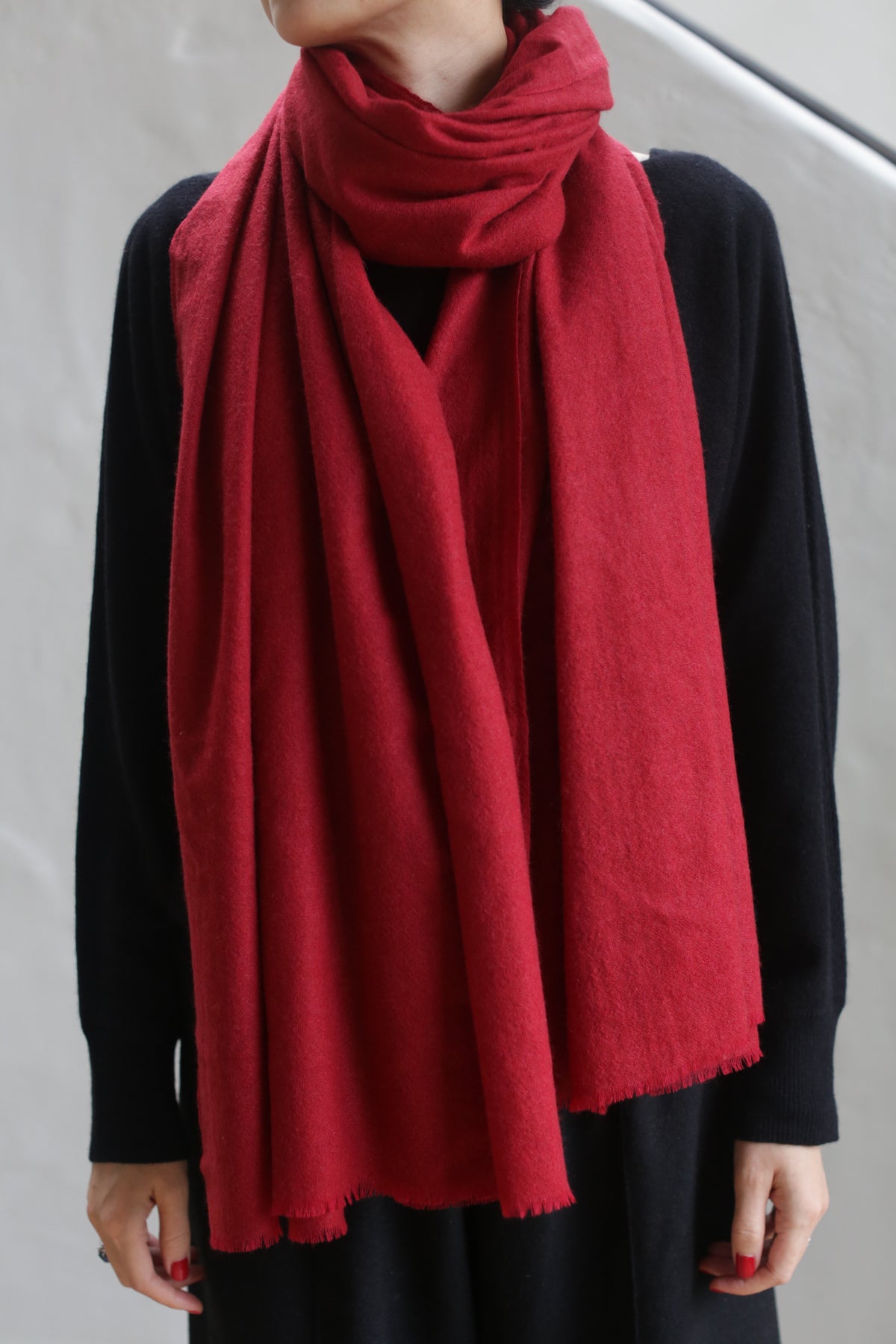 A person is wearing a black outfit with a long, Tissue Weight Cashmere Scarf in red draped around their neck. The 100% woven cashmere scarf, measuring 36" x 82", appears to be made of a soft, thick fabric and hangs down in front. The person's face is not visible in the image.
