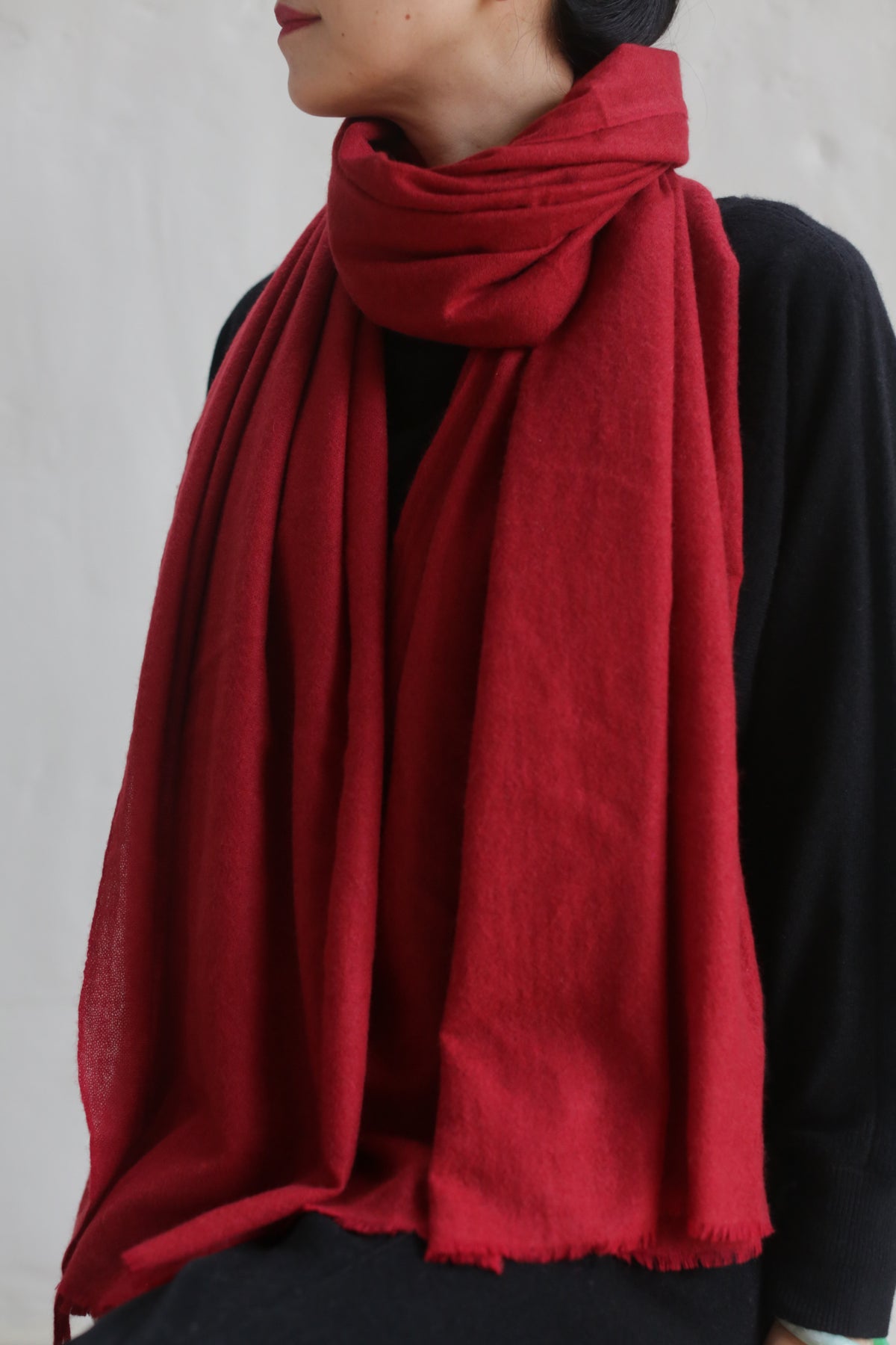 A person wearing a black outfit and a large, 100% woven Tissue Weight Cashmere Scarf in red wrapped around their neck and draping down the front. The luxurious scarf, measuring 36" x 82", stands out against the plain white background.