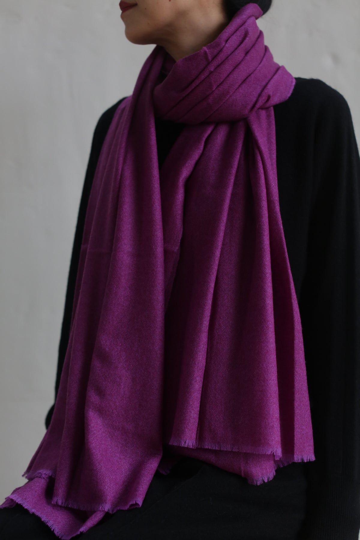 A person with short hair is wearing a dark outfit, paired with the Tissue Weight Cashmere Scarf in rich purple, wrapped elegantly around their neck. The background is plain and neutral, emphasizing the 36" x 82" scarf as the standout element of their attire.