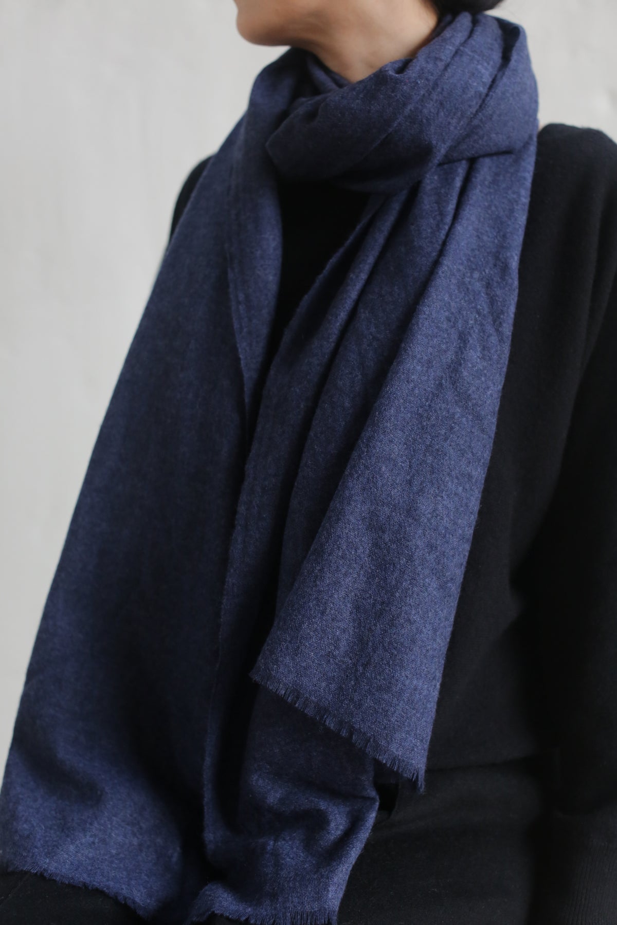 A person wearing an indigo Tissue Weight Cashmere Scarf over a black top. The 100% woven cashmere scarf, measuring 36" x 82", is wrapped around their neck, hanging down in front. The background is plain and light-colored, possibly a wall. The image focuses on the scarf and the upper torso of the person.