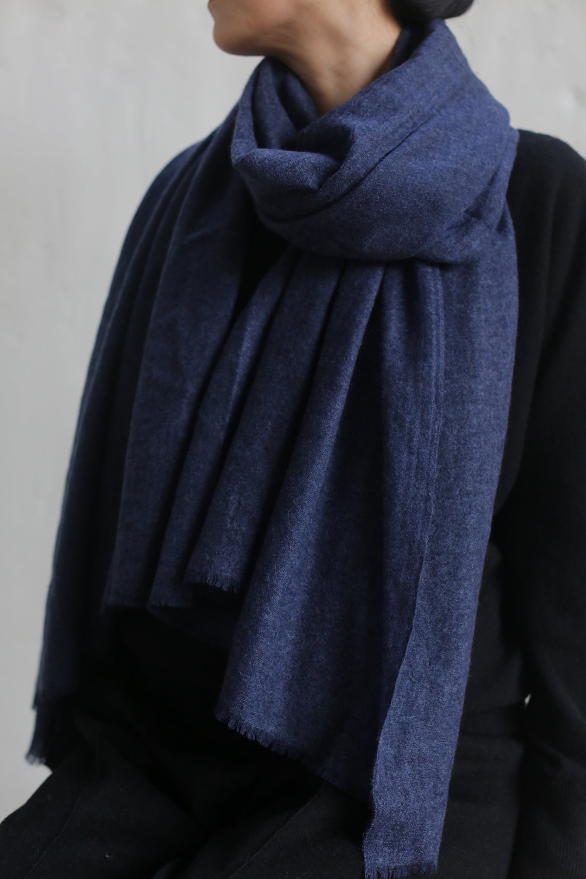A person wearing an indigo Tissue Weight Cashmere Scarf wrapped around their neck. The 36" x 82" woven cashmere scarf appears soft and is draped loosely, covering part of their chest. The background is plain and light-colored, and the person's clothing is dark.