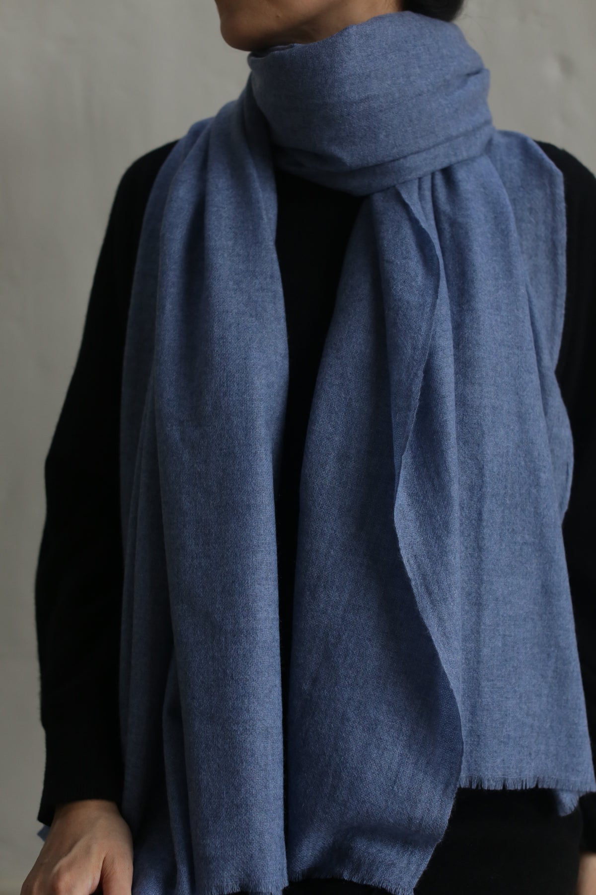 A person wearing a dark long-sleeve garment and a large, Tissue Weight Cashmere Scarf in Blue wrapped around their neck. The background is plain and neutral, focusing attention on the person and their clothing.