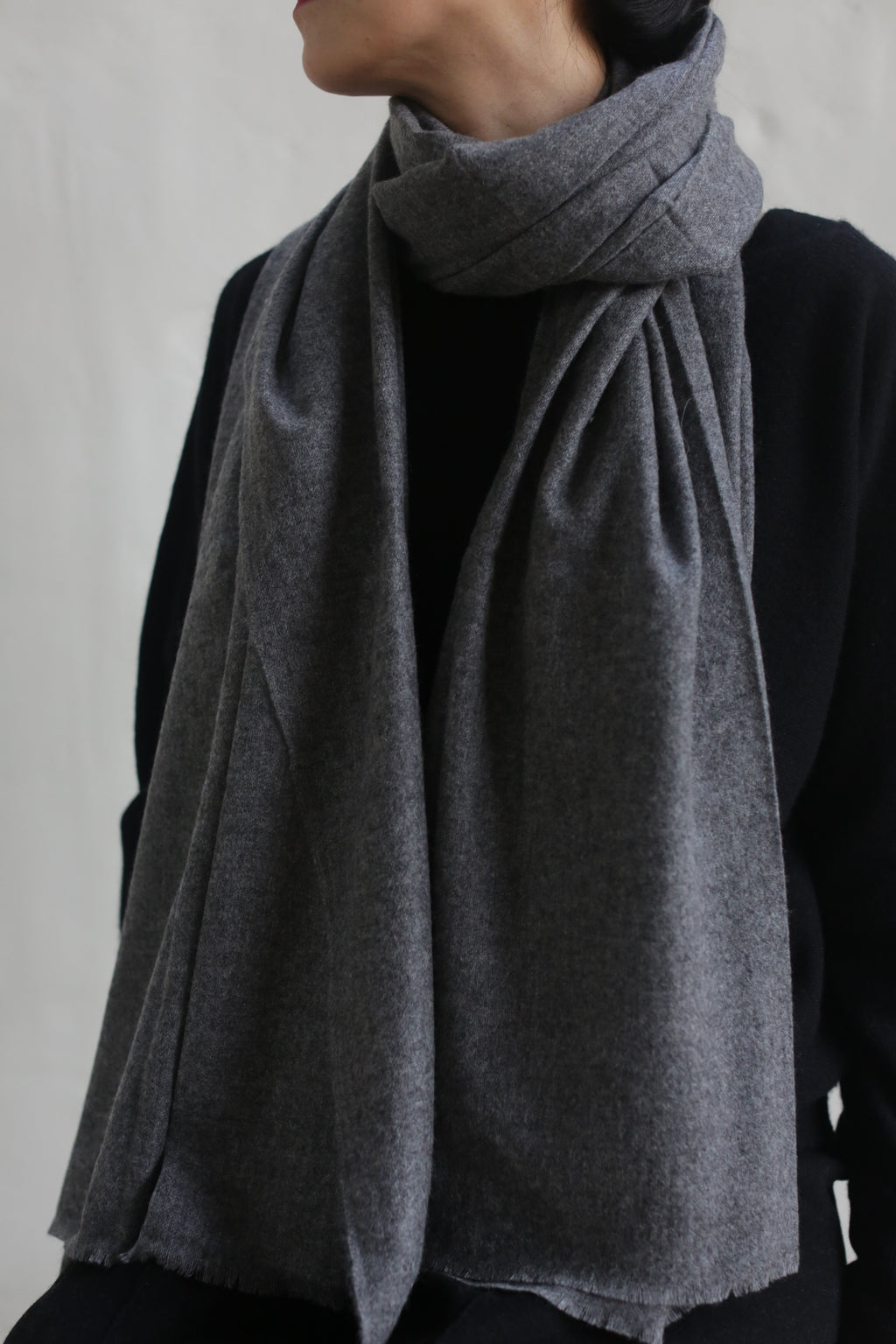 A person dressed in dark attire is wearing a Tissue Weight Cashmere Scarf in Grey, wrapped snugly around their neck with the ends draped over the front of their chest. The individual is turned slightly to the side, with their face partially out of frame against a neutral-colored background.