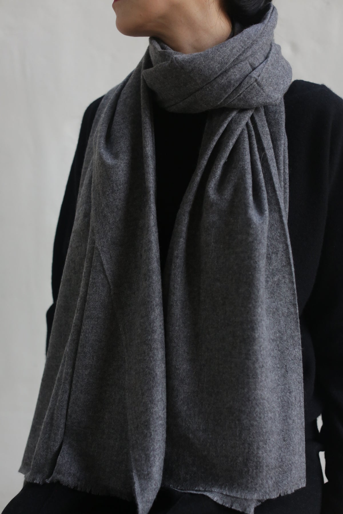 A person dressed in dark attire is wearing a Tissue Weight Cashmere Scarf in Grey, wrapped snugly around their neck with the ends draped over the front of their chest. The individual is turned slightly to the side, with their face partially out of frame against a neutral-colored background.