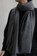 A person wearing a grey Tissue Weight Cashmere Scarf, woven from soft cashmere and draped loosely around their neck. They are dressed in a black, long-sleeved top. The background is plain and neutral, emphasizing the scarf and attire. The view is cropped to reveal only the torso and part of the neck.