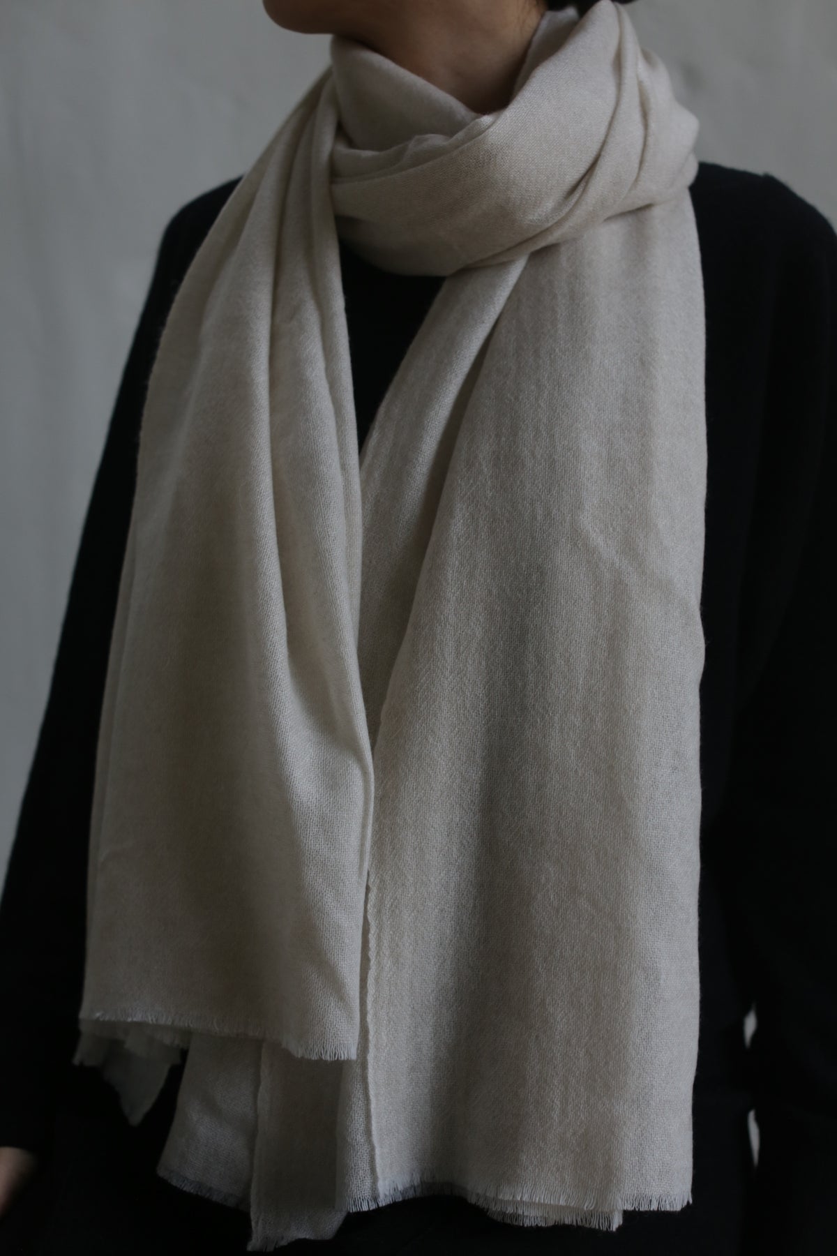 A person wearing a black top is shown from the shoulders down, with a Tissue Weight Cashmere Scarf in Oat, measuring 36" x 82", wrapped around their neck. The scarf drapes elegantly over their chest, contrasting beautifully with the dark garment. The background is neutral.