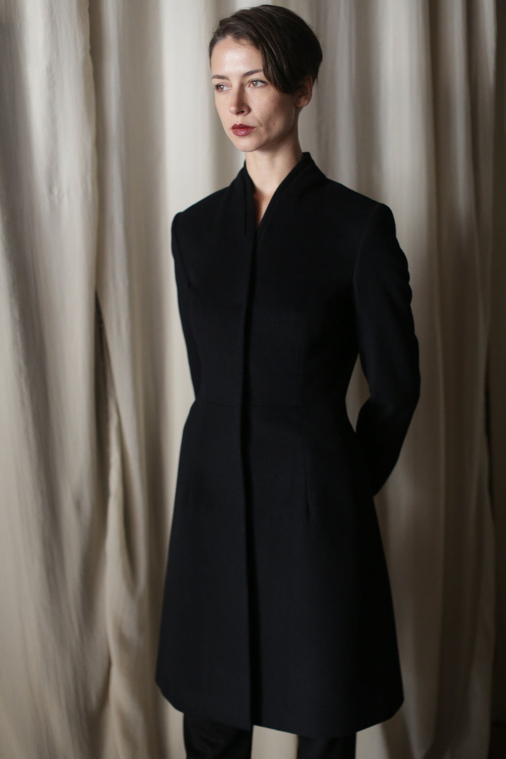 A person with short dark hair stands before cream-colored curtains, wearing the Savile | Cashmere Navy coat—a fitted, minimalist design with a high collar and their hands behind their back.