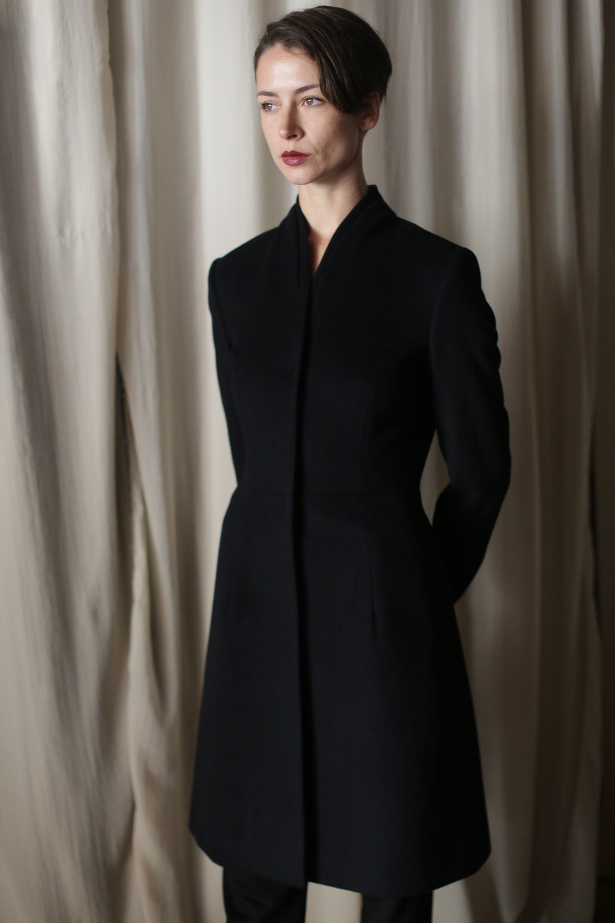 A person with short dark hair stands before cream-colored curtains, wearing the Savile | Cashmere Navy coat—a fitted, minimalist design with a high collar and their hands behind their back.