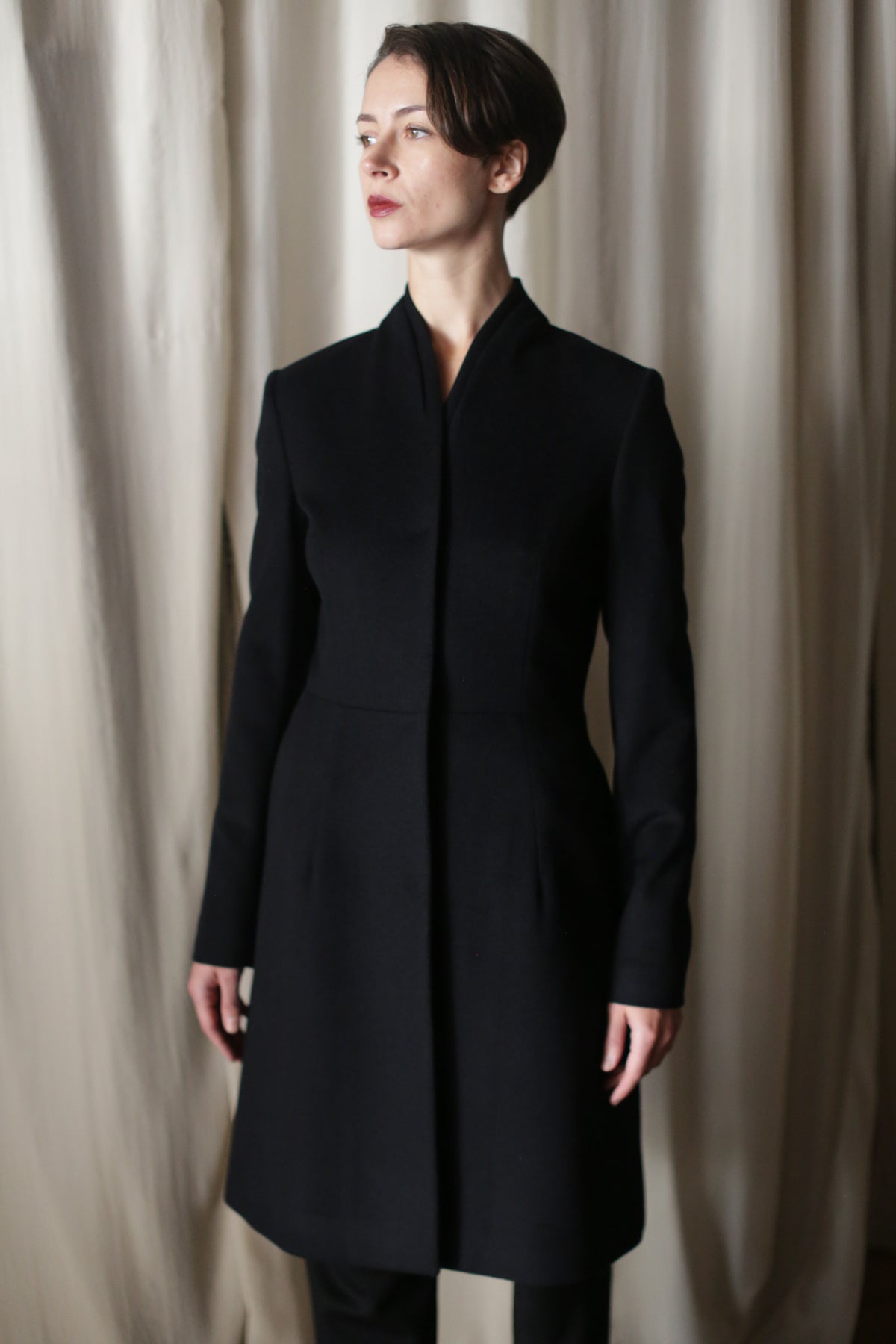 A person with short brown hair stands before light curtains, wearing the Savile | Cashmere Navy coat with a high collar, gazing slightly to the side.