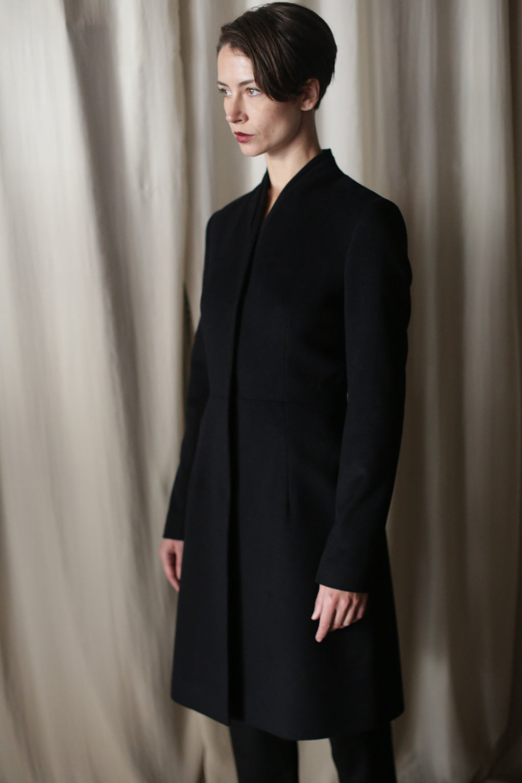 A person with short brown hair stands before beige curtains, wearing the Savile | Cashmere Navy long luxury coat. They are turned slightly to the side with a neutral expression.