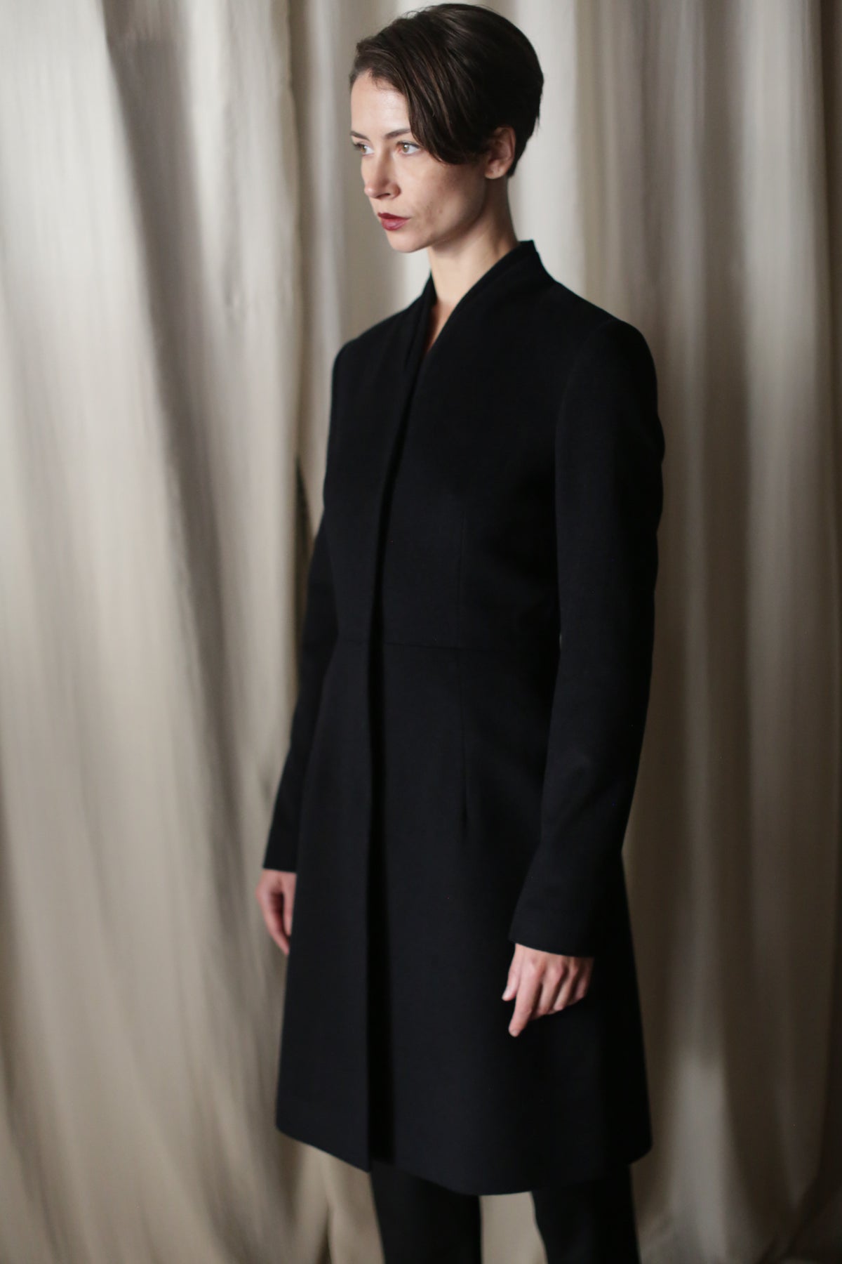 A person with short brown hair stands before beige curtains, wearing the Savile | Cashmere Navy long luxury coat. They are turned slightly to the side with a neutral expression.