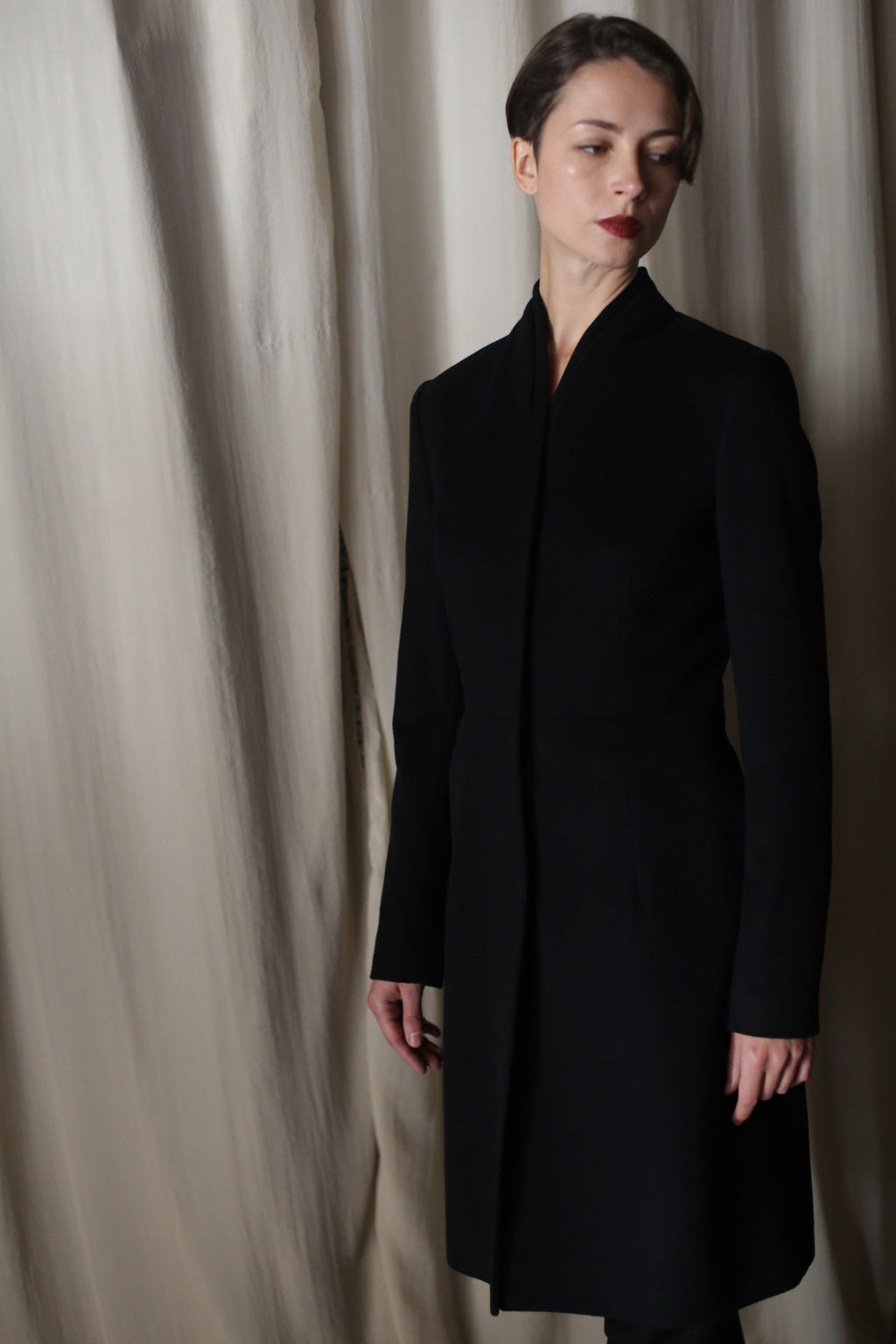 A woman with short hair wears the Savile | Cashmere Navy long minimalist coat, standing before beige curtains and gazing slightly left with a neutral expression.