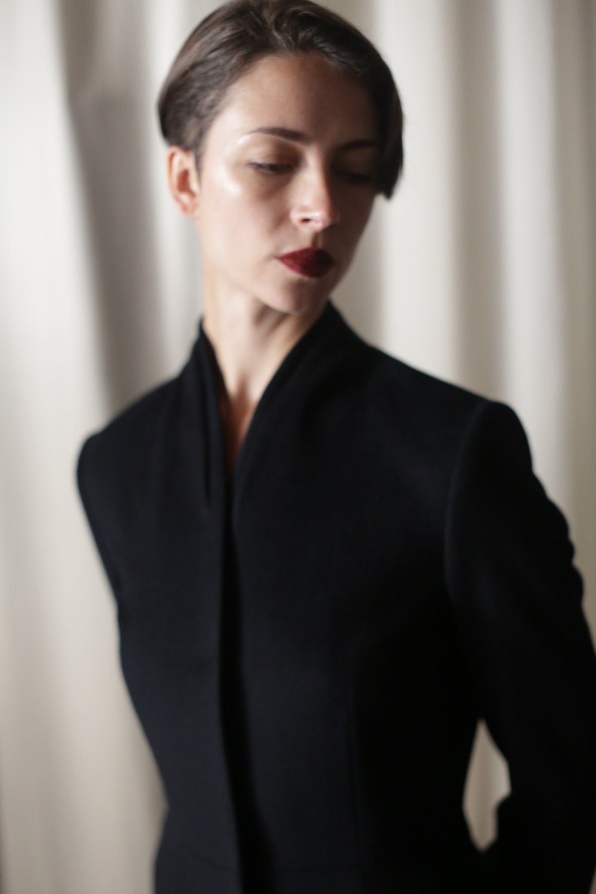 A woman with short dark hair wears the Savile | Cashmere Navy coat and red lipstick, standing against a softly blurred white curtain, looking downward with a calm expression.