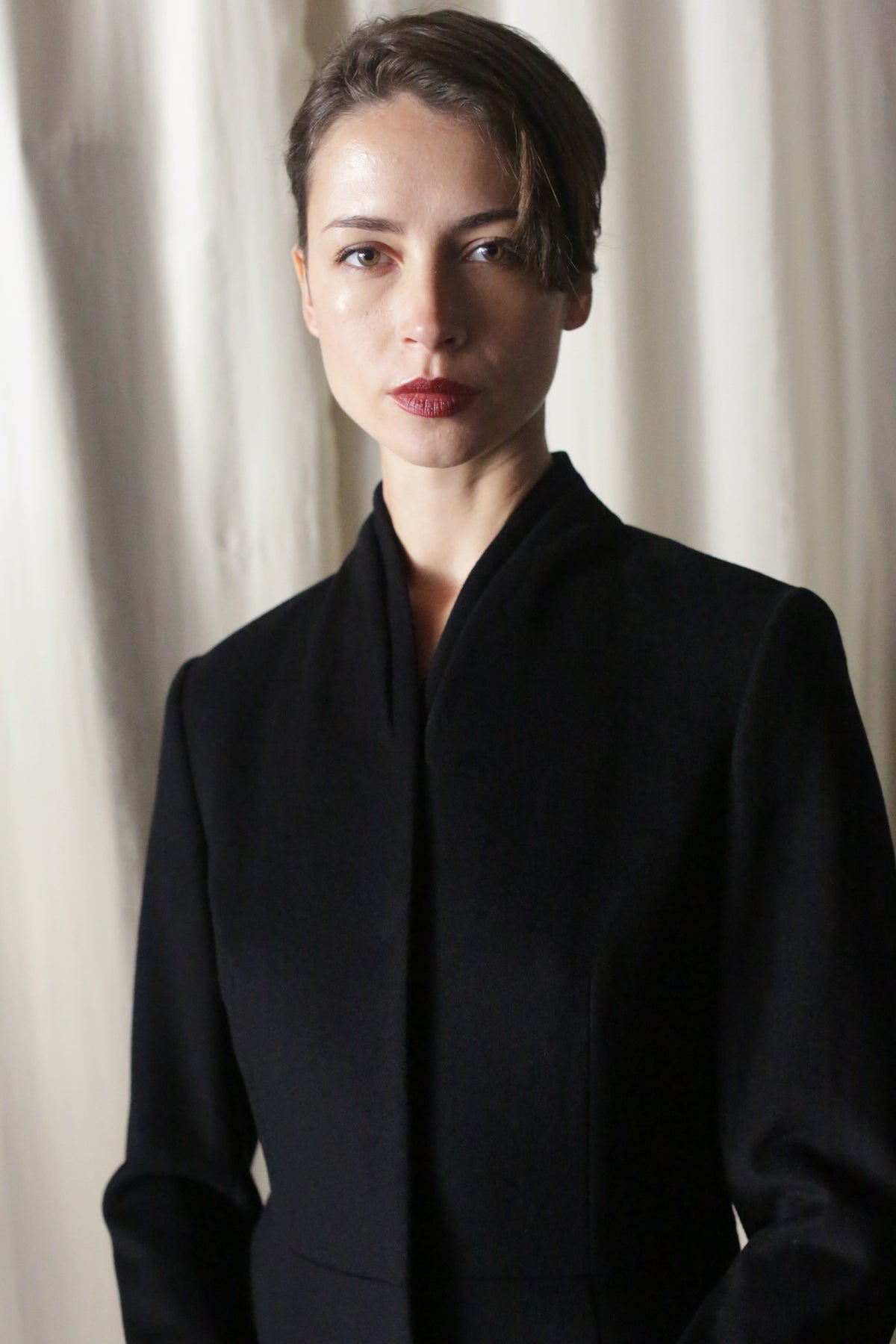 A person with short brown hair, wearing the Savile Cashmere Navy high-collared luxury coat, stands before light-colored curtains, looking directly at the camera with a neutral expression.