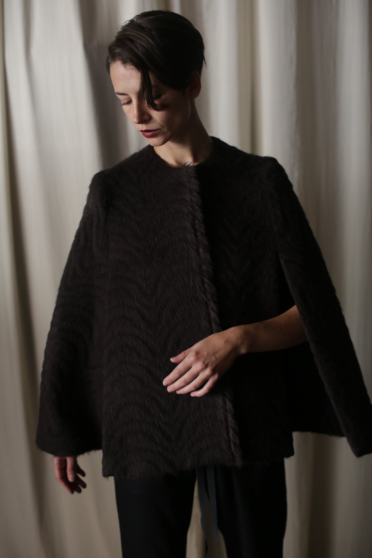 A person with short, dark hair models the Alpaca Cape, crafted from luxurious Italian alpaca, against light curtains. Wearing black pants, they gaze downward with a raised hand, capturing sustainable luxury's essence.