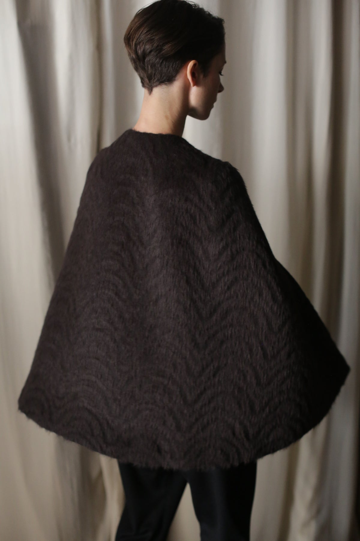 A person with short hair is seen from the back wearing an Alpaca Cape, crafted from luxurious Italian alpaca fabric. Its sustainable elegance is highlighted by light-colored curtains in the background, creating a striking contrast against this textured masterpiece.