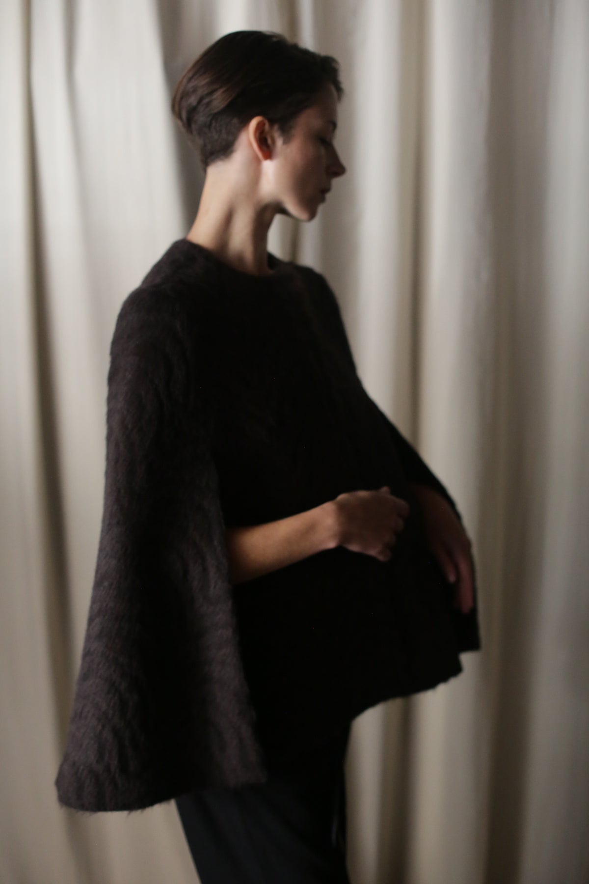 A person with short hair stands in profile against a softly lit draped curtain, wearing an Alpaca Cape with wide sleeves. Made from sustainable luxury fibers, their posture is gentle, conveying a calm and reflective mood.