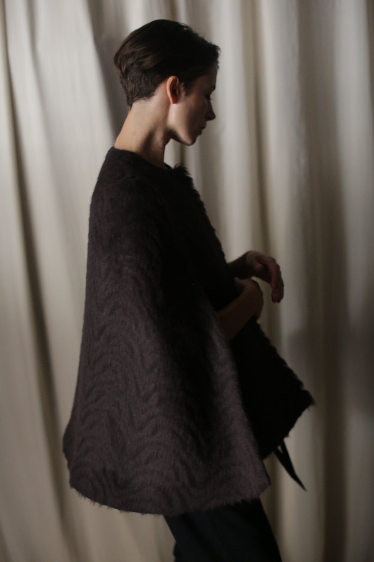 A person with short hair is seen sideways, wrapped in an Alpaca Cape made from dark Italian alpaca. The softly draped background fabric and dim lighting create a serene atmosphere, embodying sustainable luxury.