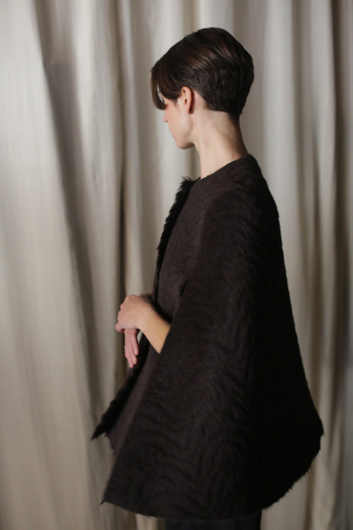 A person with short hair stands against a soft, light curtain backdrop wearing an Italian Alpaca Cape, turned to the side and looking down with hands gently clasped in a moment of sustainable luxury.