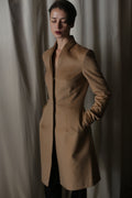 Standing with hands in pockets, the person wears the Savile | Cashmere Camel coat. Their short dark hair and closed eyes enhance the minimalist aesthetic against a soft, neutral backdrop.