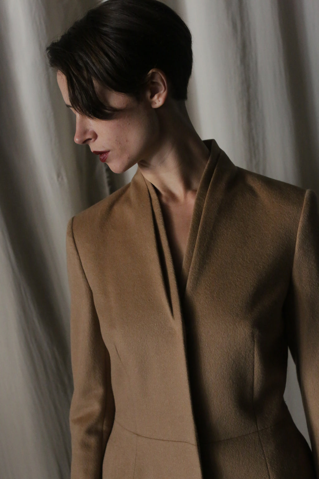 A person with short dark hair wears the Savile Cashmere Camel coat, standing against a softly lit, draped background. Their downward gaze complements the minimalist aesthetic.