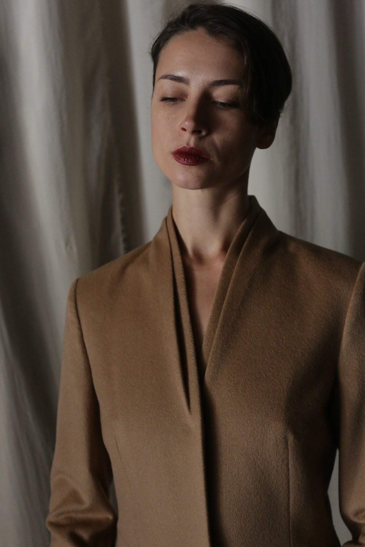 The woman with short dark hair wears a luxurious Savile | Cashmere Camel coat, eyes closed against a backdrop of draped fabric, exuding minimalist elegance.