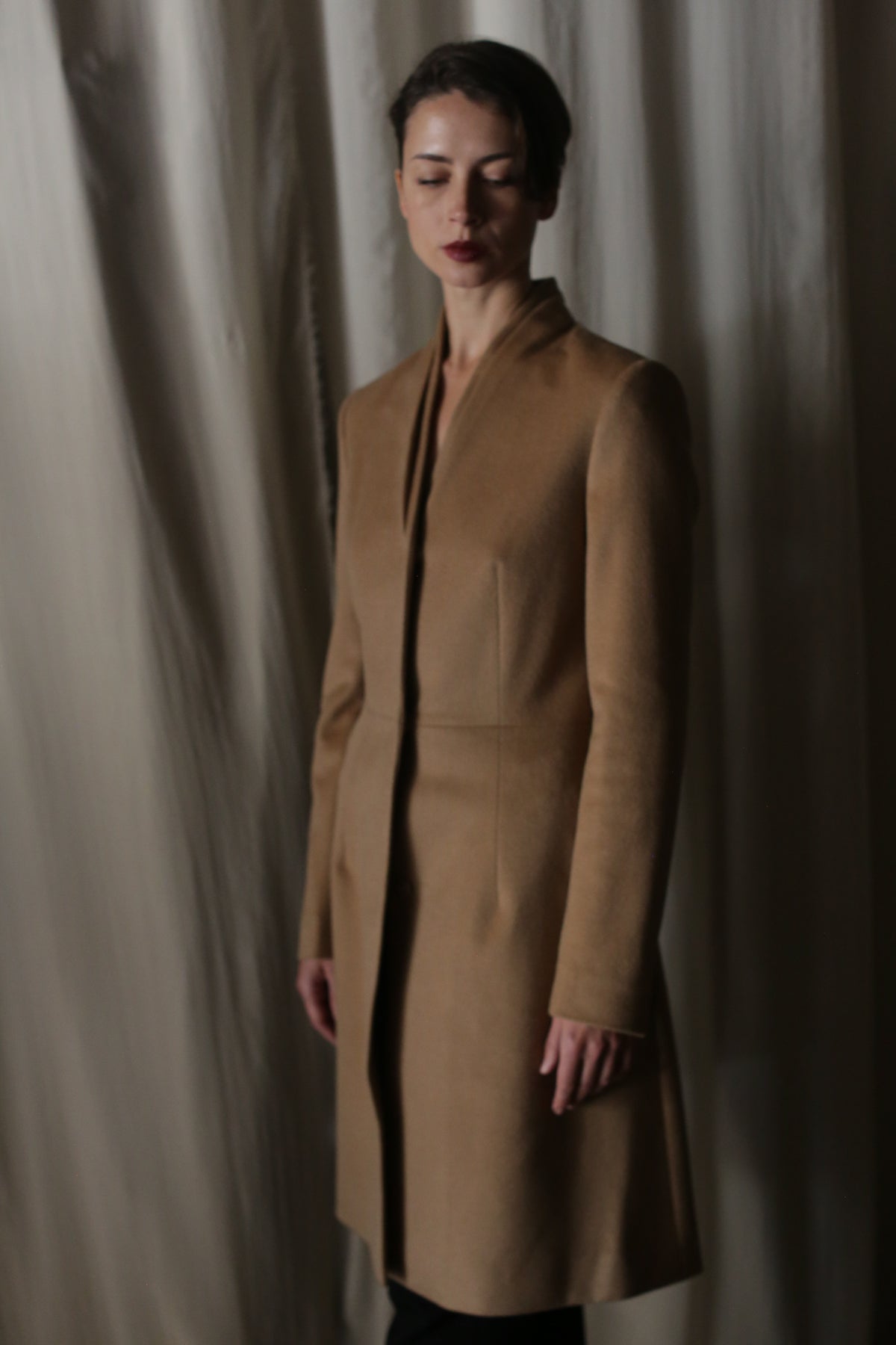 A person stands against a softly lit beige curtain, draped in the Savile Cashmere Camel—a luxurious fashion statement featuring a stylish long brown cashmere coat. With short dark hair and a minimalist aesthetic, they glance downward, exuding a serene, contemplative expression.
