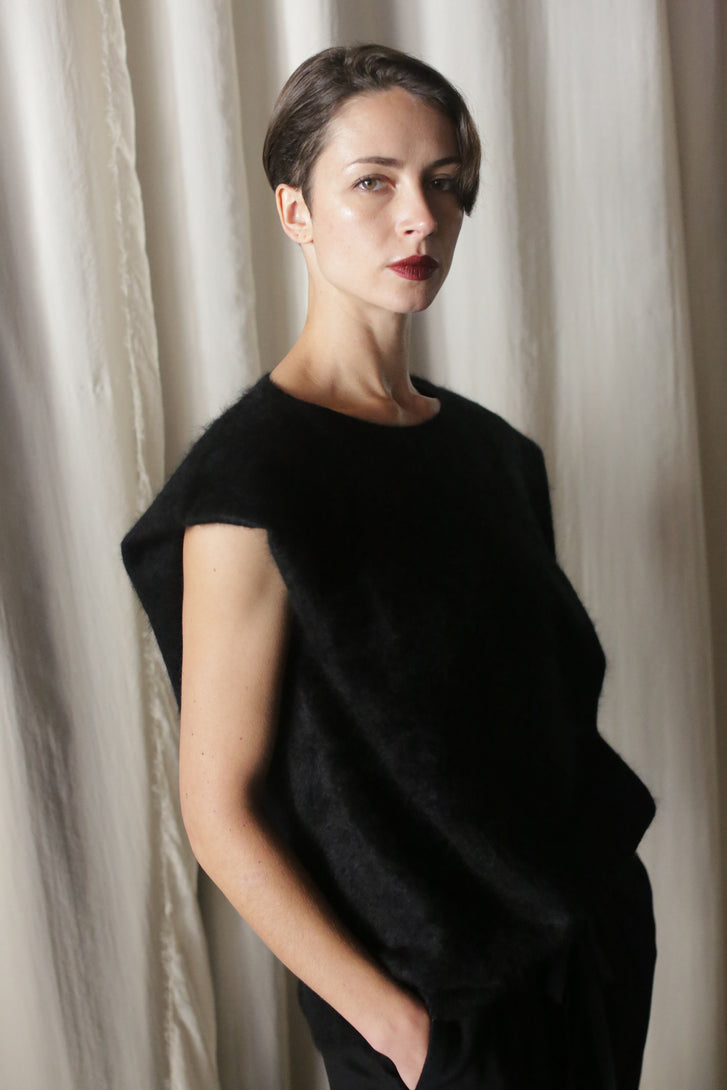 A person with short hair stands before a light curtain, wearing the Salento | Black vest over a sleeveless black top. They wear dark lipstick and maintain a neutral expression, with their left hand casually in their pocket.