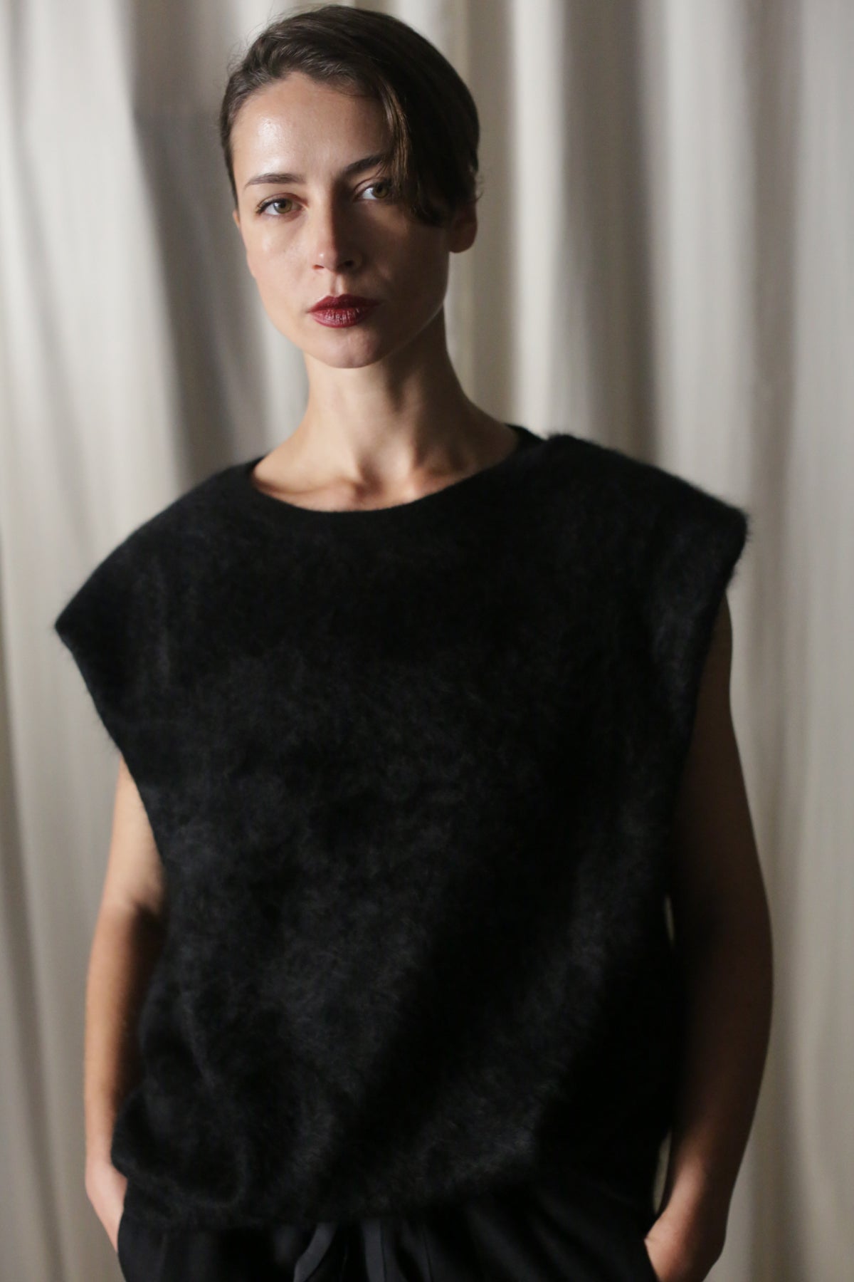 In front of a neutral curtain backdrop, a person with short, dark hair wears the stylish Salento | Black structured vest. They look calm as their hands rest casually in their pockets.