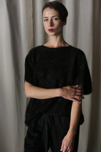 A person with short hair stands against a neutral curtain backdrop, wearing the Salento | Black structured vest over a black top and pants. One arm is relaxed by their side, while the other is bent at the elbow, resting on their opposite arm.