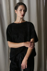 A person with short hair stands in front of a light curtain, wearing the Salento blouse and pants in black. One arm is crossed over their chest as they look directly at the camera with a neutral expression.