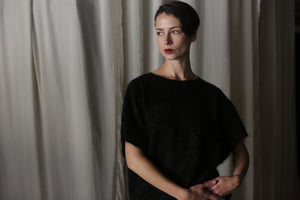 A person stands in front of beige curtains wearing the Salento | Black short-sleeved blouse. With short dark hair and a thoughtful expression, they gaze sideways as the soft lighting casts shadows, enhancing their elegant look.