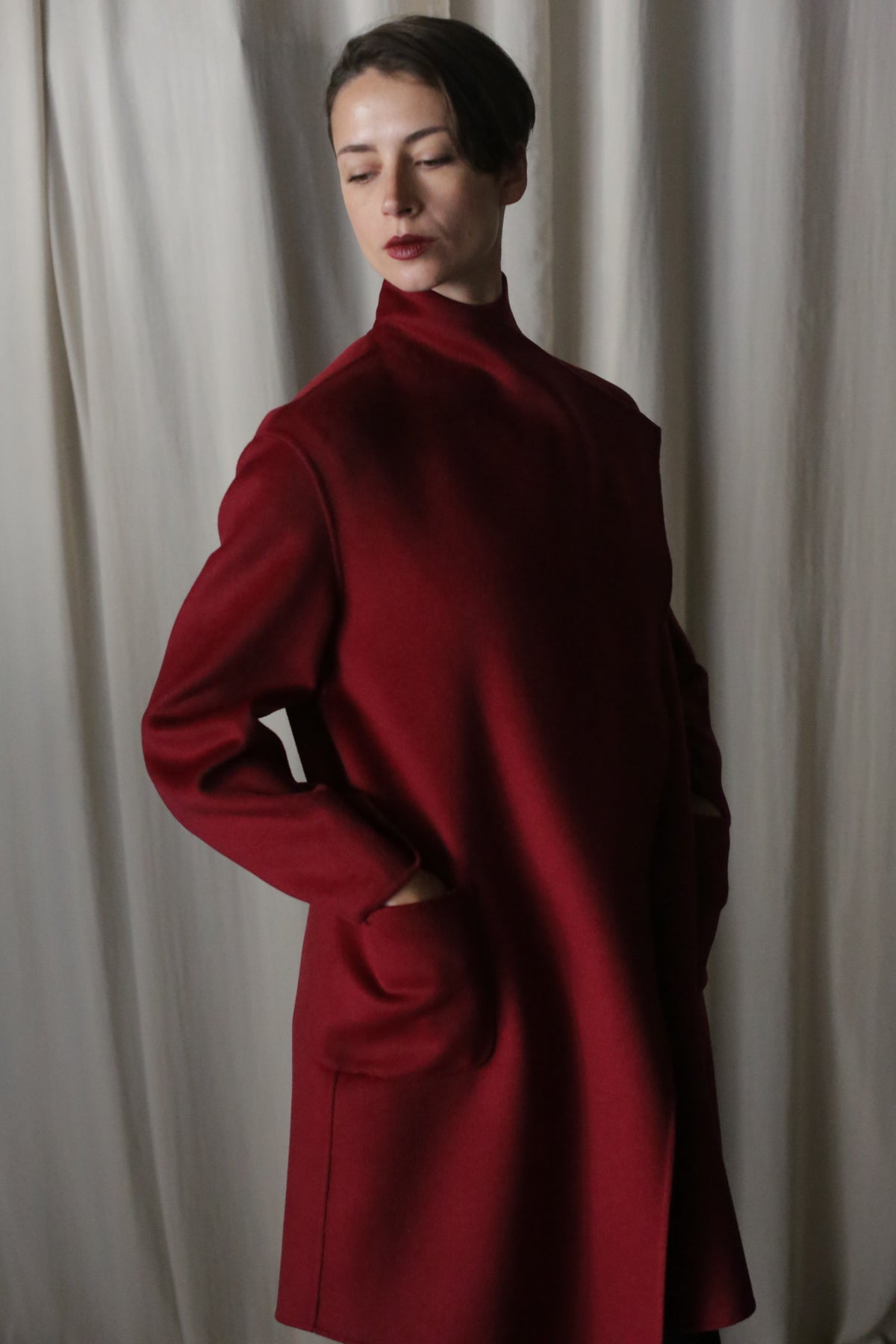 A person with short dark hair poses in the Winston Long Double-face | Wine coat against cream curtains. Made from luxurious double-faced cashmere, it boasts a draped collar and versatile style. They wear a serene expression, gazing slightly downward.