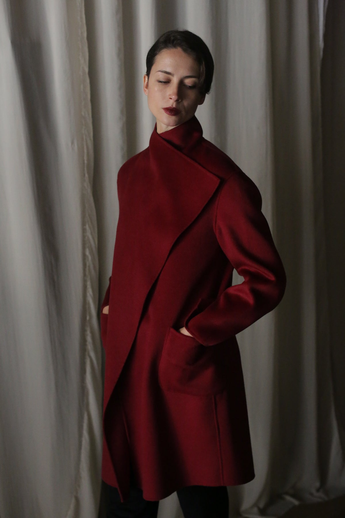 A woman in the Winston Long Double-face coat in wine, with draped collar and pockets, stands against a backdrop of softly draped fabric. Her short dark hair is styled neatly as she gazes downward serenely.