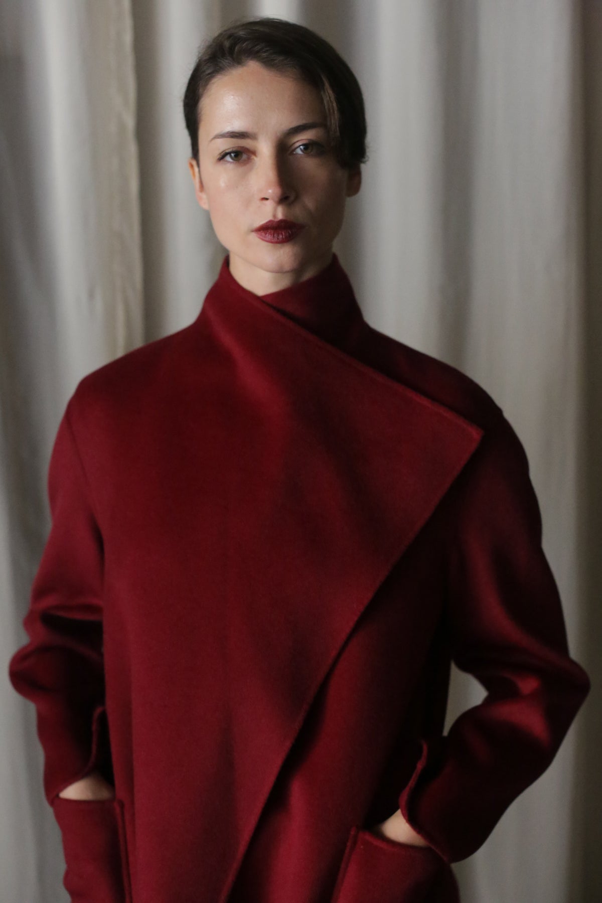 Standing against a neutral background, a person wears the Winston Long Double-face coat in wine, featuring a draped collar and crafted from double-faced cashmere. Their hands rest in pockets, gaze fixes on the camera, and their hair is neatly styled.