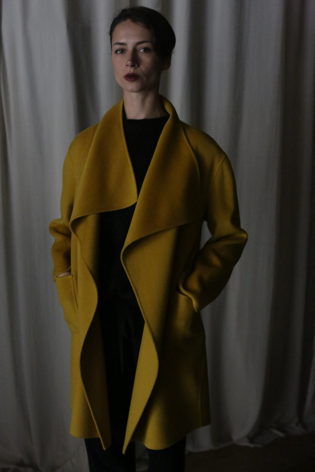 Standing confidently in a saffron Winston Long Double-face coat with hands in pockets, the draped collar accentuates its elegance. Set against a curtain backdrop, dim lighting casts soft shadows, enhancing the dramatic ambiance.
