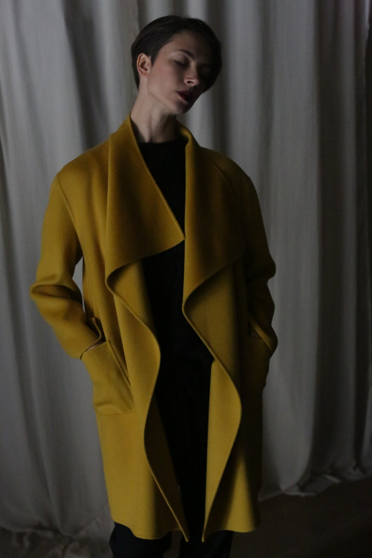A person stands in a dimly lit room wearing a Saffron Winston Long Double-face coat with large lapels. Their hands in their pockets, they are positioned against a backdrop of soft, flowing white fabric.