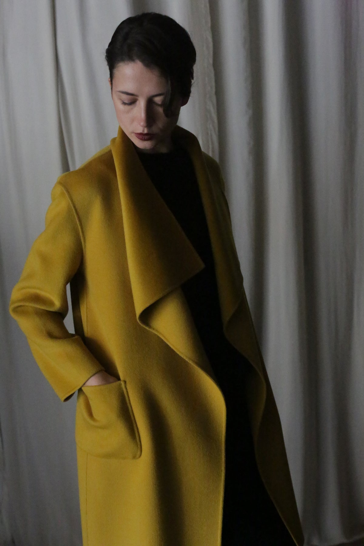 A person in a saffron Winston Long Double-face coat with hands in pockets, gazing downward against a soft-lit, neutral curtain backdrop.
