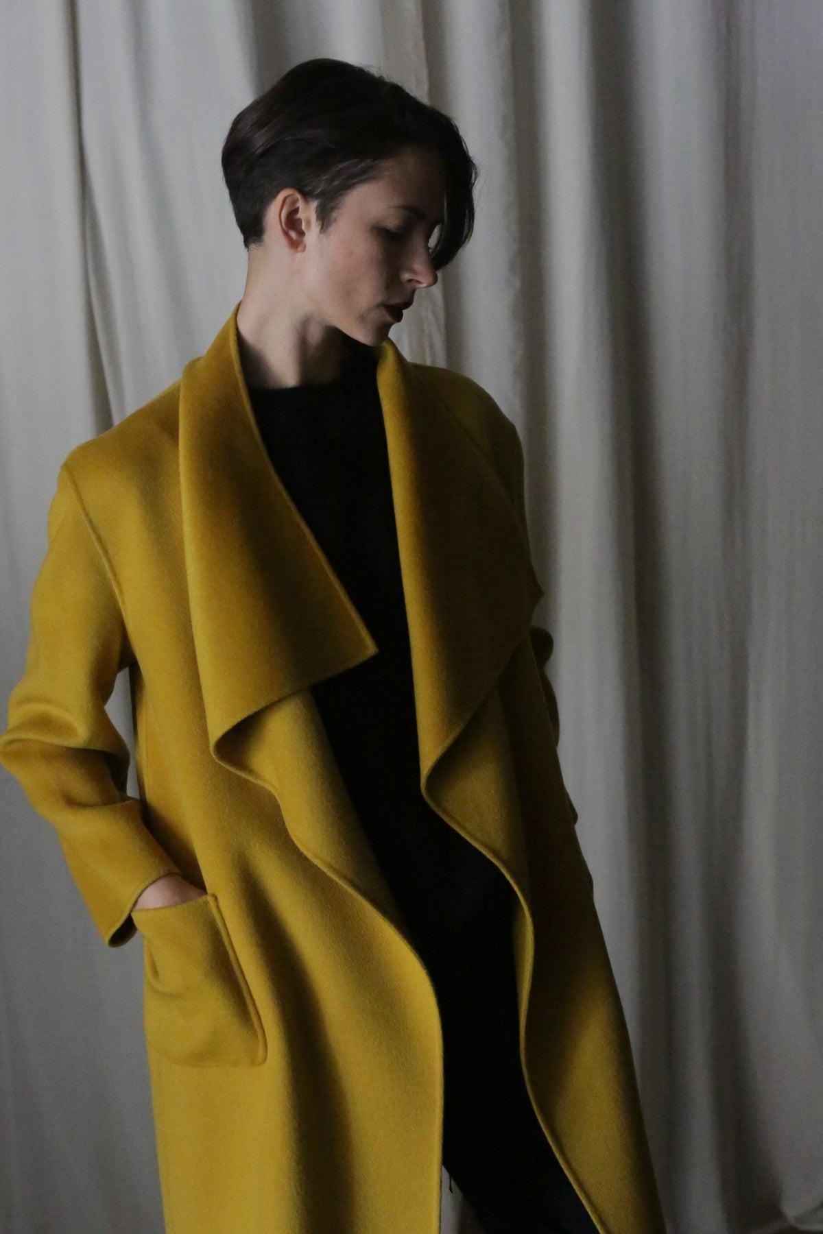 Wearing the "Winston Long Double-face" in Saffron, the individual stands against neutral drapes. The coat features a draped collar and is made from double-faced cashmere. With one hand casually resting in a pocket, they gaze downward.