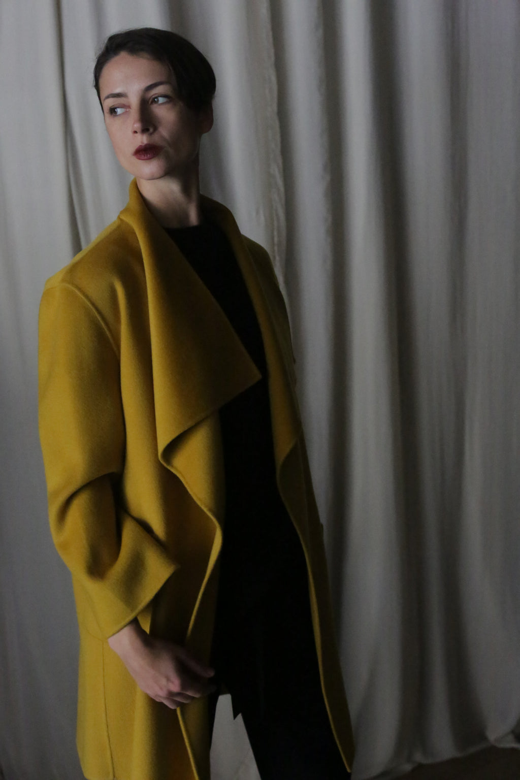 A dark-haired individual wears a Saffron-colored Winston Long Double-Face coat over a black outfit. They gaze left with a neutral expression, as white curtains softly drape in the background, enhancing the dim, moody atmosphere.