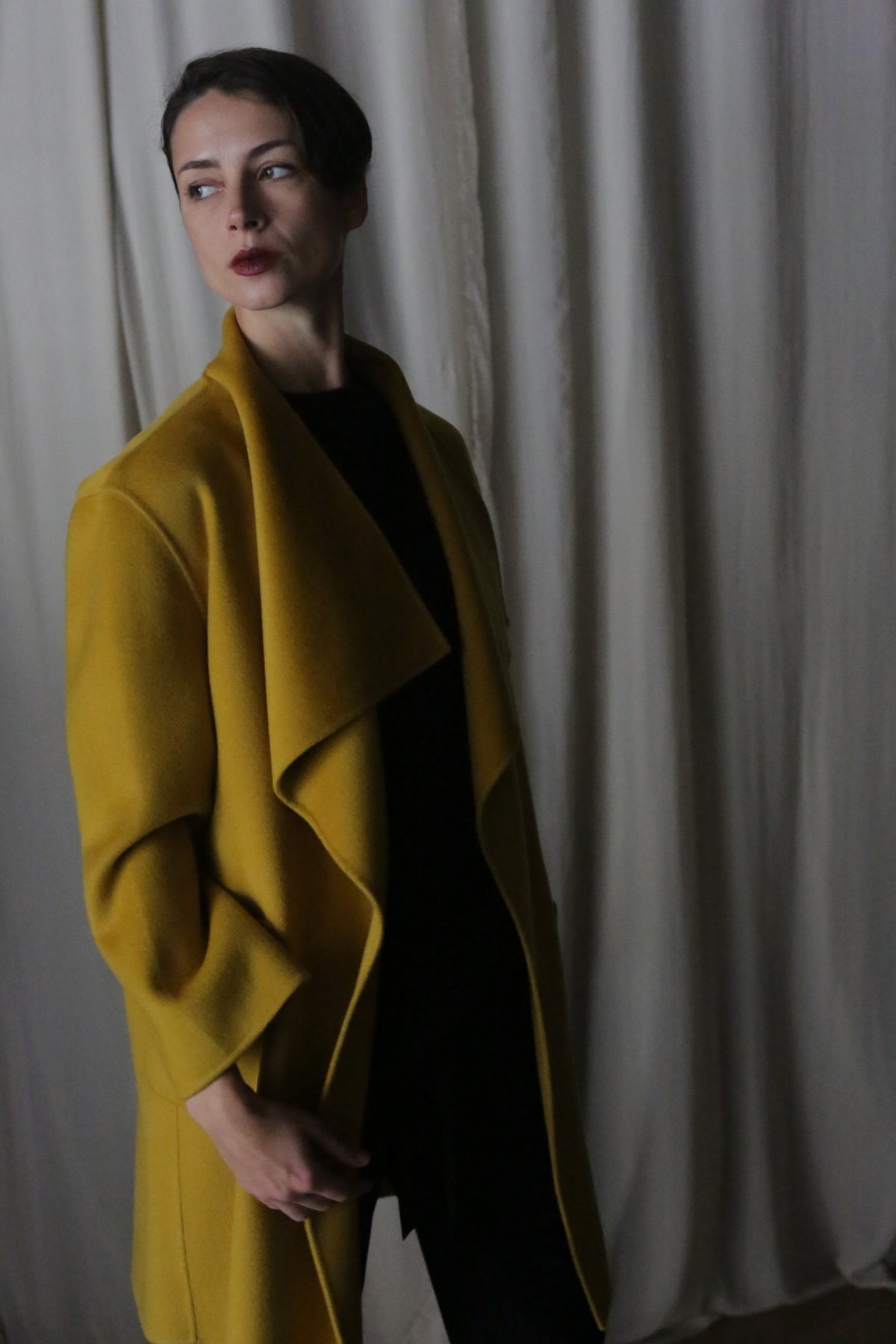 A dark-haired individual wears a Saffron-colored Winston Long Double-Face coat over a black outfit. They gaze left with a neutral expression, as white curtains softly drape in the background, enhancing the dim, moody atmosphere.