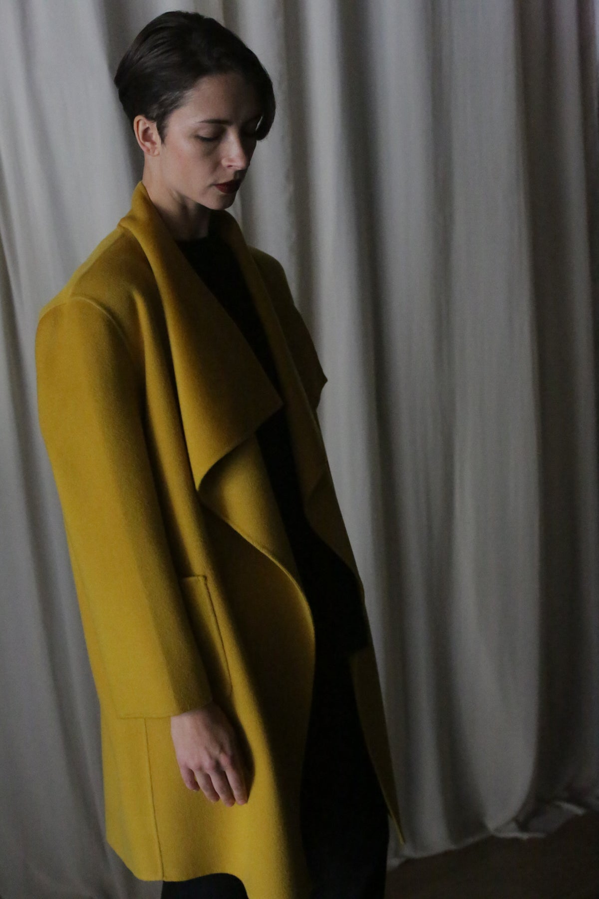 A short-haired person stands in front of draped curtains, donning a saffron Winston Long Double-Face coat over a black ensemble. The dim lighting enhances the mood and the coat's luxurious cashmere.