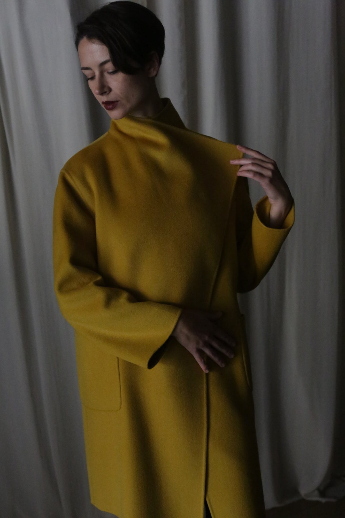 A person with short dark hair poses serenely against a curtain, wearing the Winston Long Double-face coat in Saffron. With closed eyes and a tranquil expression, they gently touch its draped collar. The soft lighting adds to the subdued atmosphere.