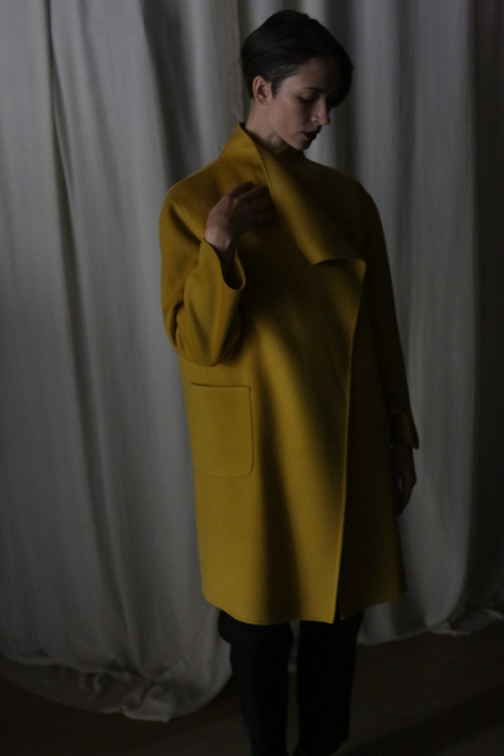 In a dimly lit room with curtain backdrops, a person stands enveloped in a dramatic atmosphere wearing the Winston Long Double-face coat in Saffron. The cashmere coat features a draped collar and two visible pockets, highlighted by soft shadows cast by the lighting.
