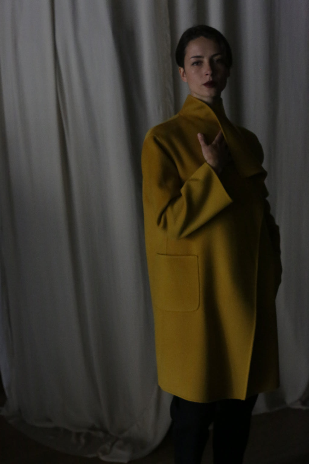 In a dimly lit room, someone leans against soft, white fabric wearing the Winston Long Double-face coat in Saffron. The draped collar and gentle shadows add an air of elegance and mystery to the moody ambiance.