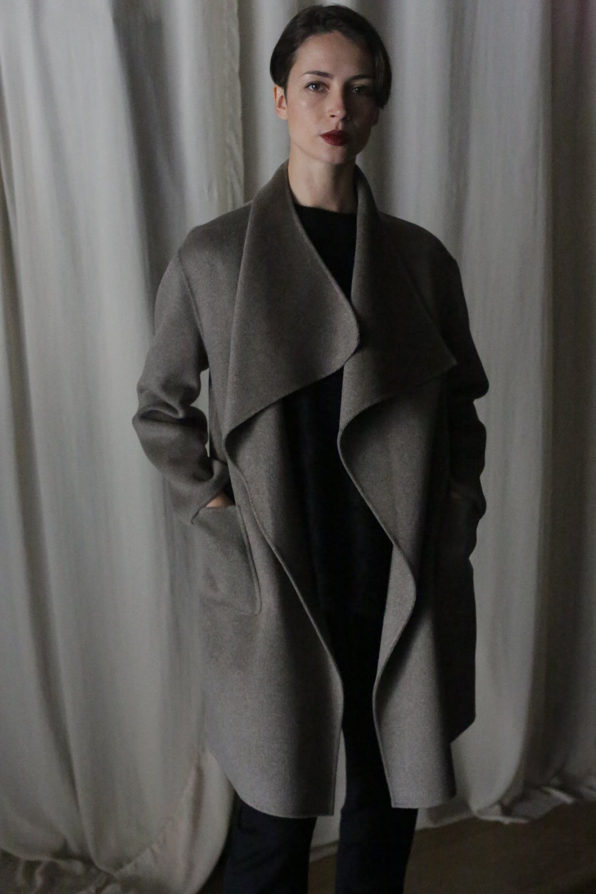A person stands confidently with hands in pockets, wearing the Winston Long Double-face coat in Undyed Natural and dark pants, embodying timeless neutrals. Their hair is short, and they wear dark lipstick against a softly lit curtain backdrop.
